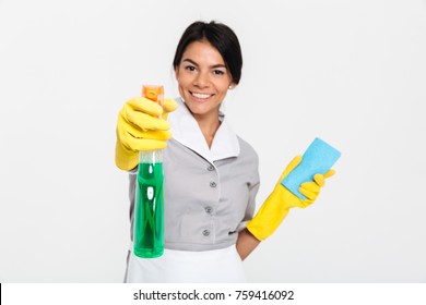 Close-up Portrait Of Professional Housekeeper In Uniform And Yellow Rubber Gloves Spraying The Cleaner On You, Isolated On White Background