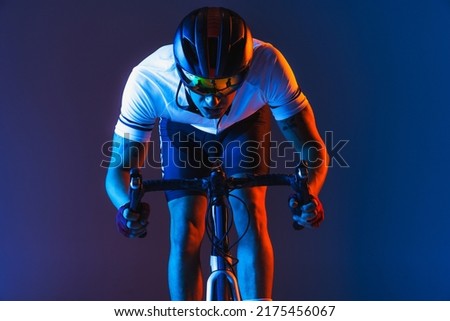 Closeup portrait of professional male cyclist in sports uniform, goggles and helmet on blue background in neon. Concept of active life, rest, travel, energy, sport. Copy space for ad. Front view