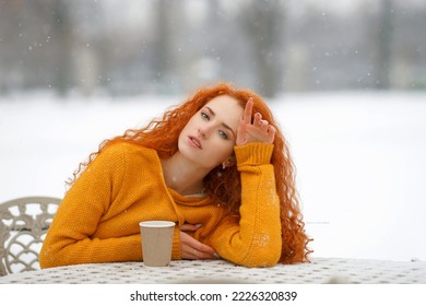                Close-up portrait of a red-haired beautiful girl looking at the camera sitting at the table on the street in winter, it's snowing hot with coffee Cold winter,
 white snowflake  - Powered by Shutterstock