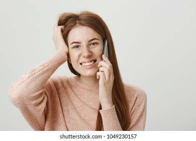 Close-up Portrait Of Redhead Girl With Freckles Being Troubled While Speaking On Phone, Scratching Head And Smiling At Camera, Standing Against Gray Background. Woman Do Not Know What She Should Say