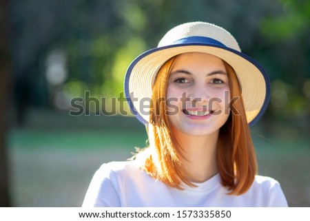Closeup portrait of redhead hipster teenage girl in yellow hat smiling outdoors in sunny summer park.