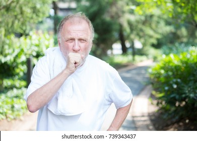 Closeup portrait, senior guy holding towel, very tired, exhausted from over exertion, coughing catching breath, isolated outdoors outside green trees background