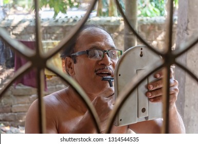 Close-up Portrait Of Senior Indian Man Shaving Beard With A Razor In Front Of A Small Mirror. 