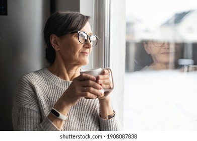 Close-up portrait of senior older woman wearing glasses enjoys morning coffee in the kitchen at home. A modern retirement lady daydreaming with a mug of hot drink looks through the window - Powered by Shutterstock