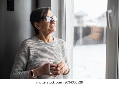 Close-up Portrait Of Senior Older Woman Wearing Glasses Enjoys Morning Coffee In The Kitchen At Home. A Modern Retirement Lady Daydreaming With A Mug Of Hot Drink Looks Through The Window