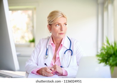 Close-up Portrait Of Senior Overwhelmed Female Doctor Sitting At Medical Room And Looking Thoughtfully While Writing Something. 