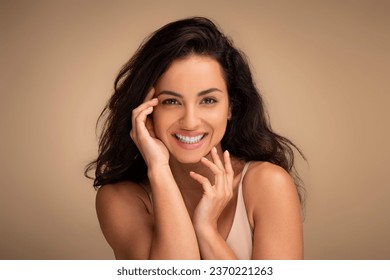 Closeup portrait of sensual beautiful young brunette long-haired woman posing on beige studio background, touching her face and smiling, enjoying her smooth glow face skin after beauty treatment