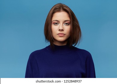 Closeup Portrait Of Serious Beautiful Young Woman With Dark Hair Wears Blue Pullover Feels Unhappy, Abused And Looks To The Camera Isolated Over Blue Background