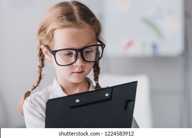 Close-up Portrait Of Serious Little Child In Eyeglasses Writing In Clipboard