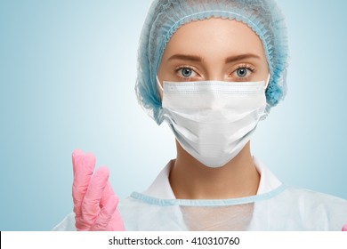 Close-up Portrait Of Serious Woman Nurse Or Doctor In White Surgical Mask, Blue Cap And Rubber Sterile Gloves Looking Confident At The Camera Isolated Against Blank Studio Background. Surgery Concept