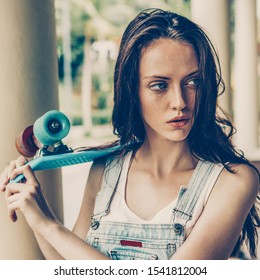 Closeup Portrait Of Sexy Beautiful Woman In White T-shirt And Short Denim Overall Holding Blue Short Skate Board. Urban Scene, City Life. Cute Attractive Sexy Hipster Lady Standing In Front Of Column.