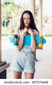 Close-up Portrait Of Sexy Beautiful Woman In White T-shirt And Short Denim Overall Holding Short Skate Board. Urban Scene, City Life. Cute Attractive Sexy Hipster Lady Standing In Front Of A Column.