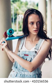 Closeup Portrait Of Sexy Beautiful Woman In White T-shirt And Short Denim Overall Holding Blue Short Skate Board. Urban Scene, City Life. Cute Attractive Sexy Hipster Lady Standing In Front Of Column.