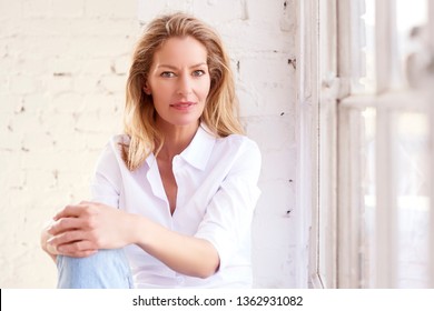 Close-up Portrait Shot Of Beautiful Blond Mature Woman Wearing White Shirt And Jeans While Relaxing By The Window.