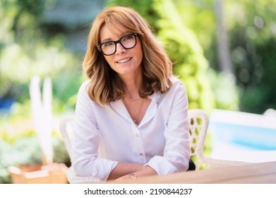 Close-up portrait shot of cheerful middle aged woman wearing glasses and white shirt while relaxing on the patio at home. - Shutterstock ID 2190844237