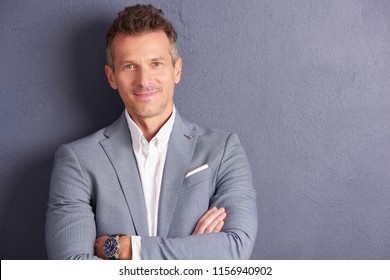 Close-up portrait shot of confident middle aged businessman with folded arms standing at grey wall and looking at camera.