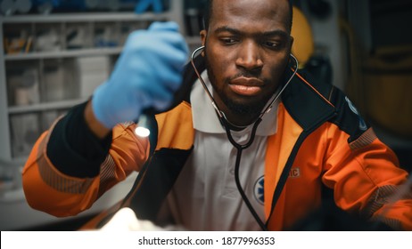 Close-up Portrait Shot Of A Serious And Focused Black African American Paramedic In An Ambulance Vehicle With An Injured Patient. Emergency Medical Technician Uses Stethoscope To Monitor The Condition