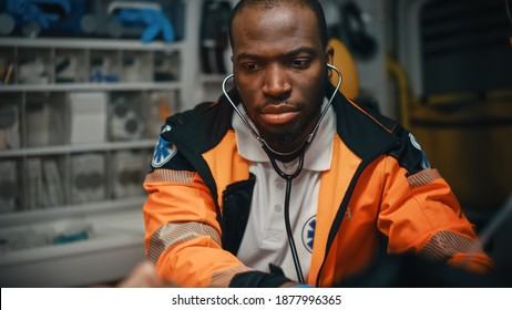 Close-up Portrait Shot Of A Serious And Focused Black African American Paramedic In An Ambulance Vehicle With An Injured Patient. Emergency Medical Technician Uses Stethoscope To Monitor The Condition