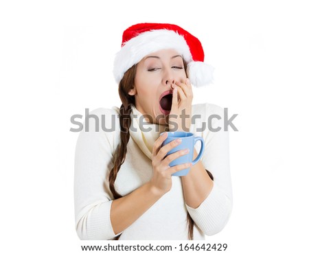 Closeup portrait of sleepy young cute christmas girl, attractive woman, tired, yawning holding cup of hot beverage, wearing Santa Claus hat, isolated on white background. Busy holiday season concept.