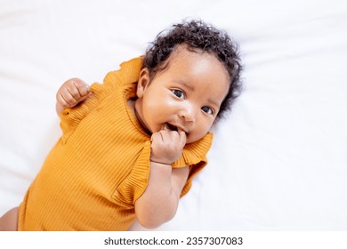 a close-up portrait of a small African-American baby girl in an orange bodysuit on the bed at home, a funny six-month-old black newborn baby lies on the back and smiles. View from above. - Powered by Shutterstock