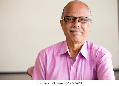 Closeup portrait, smart elderly man in pink shirt with dark eye glasses, specs, sitting down, isolated indoors white chalkboard background