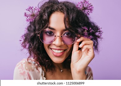 Close-up Portrait Of Smiling Brown-eyed Woman With Flowers In Black Hair. African Blissful Lady In Sunglasses Posing On Purple Background.