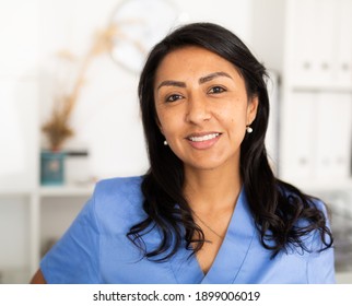 Closeup Portrait Of Smiling Hispanic Woman Health Worker Wearing Blue Uniform Standing In Medical Office