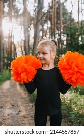 Close-up Portrait Of A Smiling Professional Sportswoman Girl, Child Cheerleader In A Black Suit With Orange Big Pom-poms In Her Hands In The Forest At Sunset. Sports Training For Cheerleading.