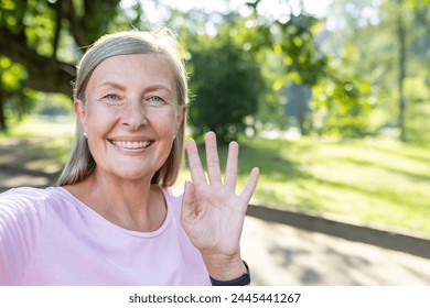 Close-up portrait of a smiling senior gray-haired woman standing outside in a park, talking and greeting on a mobile phone camera. - Powered by Shutterstock