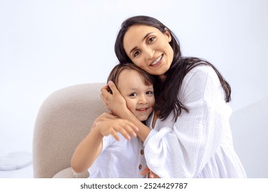 A close-up portrait of a smiling woman hugging a little boy at home. Mother and her kid in casual clothes are sitting on the couch. Embraces of a young brunette and her cute son. Minimalistic interior - Powered by Shutterstock