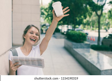 Closeup Portrait Of Smiling Young Beautiful Woman Leaning On Wall, Holding Drink And Newspaper, Raising Hand And Greeting Someone On Street. Front View.