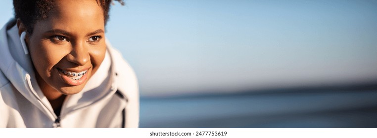 Closeup Portrait Of Smiling Young Black Lady Athlete Resting Outdoors After Training, Motivated Sporty African American Woman Wearing Wireless Earphones And Dental Braces Looking Away, Copy Space - Powered by Shutterstock