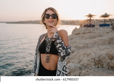 Closeup Portrait Of Smiling Young Girl On Beach, Sunset Time. Wearing Trendy Black Swimsuit, Bikini, Stylish Sunglasses, Necklace, Cardigan, Cape With Ornaments.