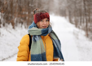 Closeup portrait of a smiling young woman wearing a red knit headband, scarf, and yellow jacket on a cold winter day. Girl dressed warmly  surrounded by a blurred snowy forest. - Powered by Shutterstock