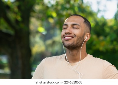 Close-up portrait of sportsman in park, hispanic man jogging in park with eyes closed breathing fresh air and resting, jogging with headphones listening to music and online radio and podcasts. - Powered by Shutterstock