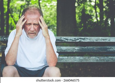 Closeup Portrait, Stressed Older Man In White Shirt, Hands On Head With Bad Headache, Sitting On Bench, Isolated Background Of Trees Outside.