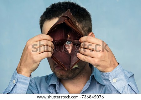 Closeup portrait, stressed, upset, sad, unhappy young man standing with, looking into hole of empty wallet. Financial difficulties, bad economy concept. Negative emotion