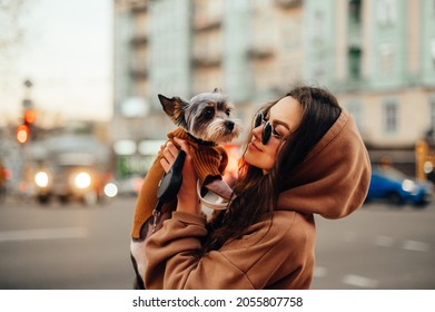 Closeup Portrait Of A Stylish Caucasian Woman Wearing Shades And Hoodie, Holding Her Her Adorable Dog - Yorkie Terrier, Standing On A City Street. Blurred Background.