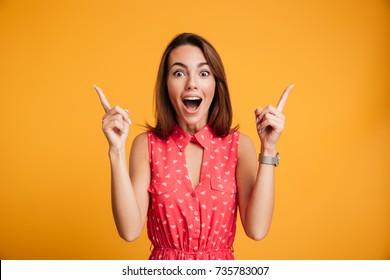 Close-up portrait of surprised pretty young woman in red dress pointing with two fingers, looking at camera with open mouth, isolated on yellow background