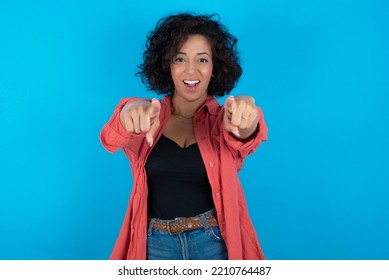 Close-up portrait of surprised young beautiful woman with curly short hair wearing red overshirt over blue wall pointing with two fingers to the camera saying: I choose you!, looking up with open mout