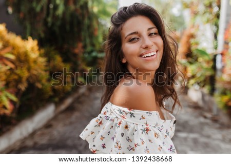 Close-up portrait of tanned woman on rest in light blouse. Curly dark-haired girl with snow-white smile looks into camera on background of tropical plants