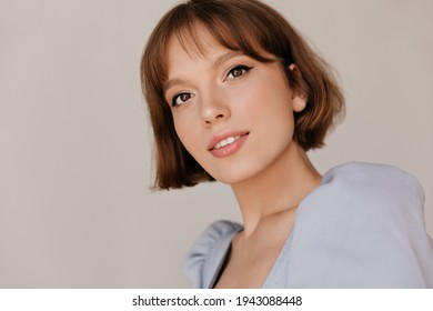 Close-up portrait of tender girl isolated over light background. Attractive young lady with pure skin, short dark hairstyle and nude makeup, wearing blue outfit, looking into camera