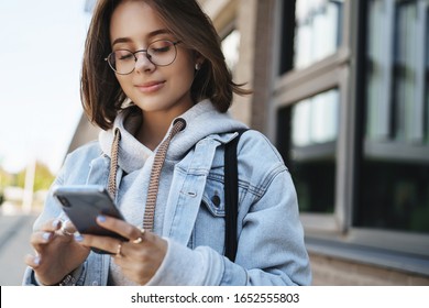 Close-up portrait tender romantic girlfriend send cheerful message, heart emoji friend, holding mobile phone and smiling at display, texting with friend, communicating while standing on street