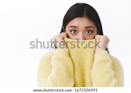 Close-up portrait timid and insecure cute asian girl pull sweater collar on face as feel afraid and scared of horror movie, eyes exress fear, trembling frightened, standing white background