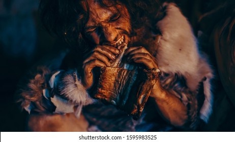 Close-up Portrait of Tribe Leader Wearing Animal Skin Eating in a Dark Scary Cave at Night. Neanderthal or Homo Sapiens Family Cooking Animal Meat over Bonfire and then Eating it.