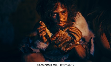 Close-up Portrait of Tribe Leader Wearing Animal Skin Eating in a Dark Scary Cave at Night. Neanderthal or Homo Sapiens Family Cooking Animal Meat over Bonfire and then Eating it.