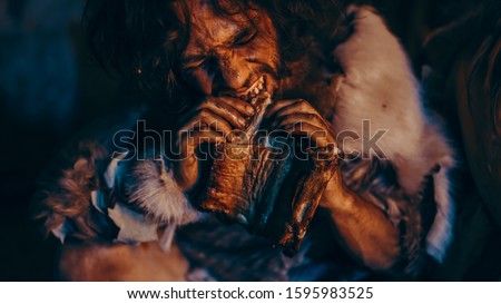 Close-up Portrait of Tribe Leader Wearing Animal Skin Eating in a Dark Scary Cave at Night. Neanderthal or Homo Sapiens Family Cooking Animal Meat over Bonfire and then Eating it.