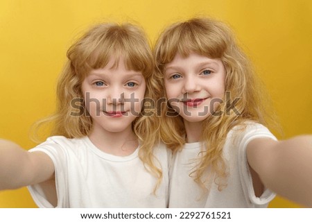 Close-up portrait of twin girls. Two beautiful and positive curly blonde sisters who are 6 years old, wearing white T-shirts, taking a selfie on a smartphone on a bright isolated yellow background