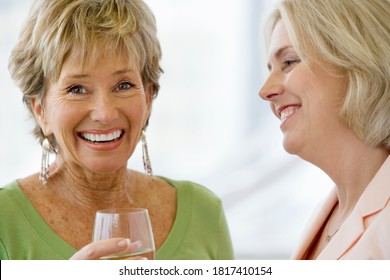 Close-up Portrait Of Two Senior Women Talking At Social Gathering Holding Wine Glasses And Laughing With One Looking At The Camera.