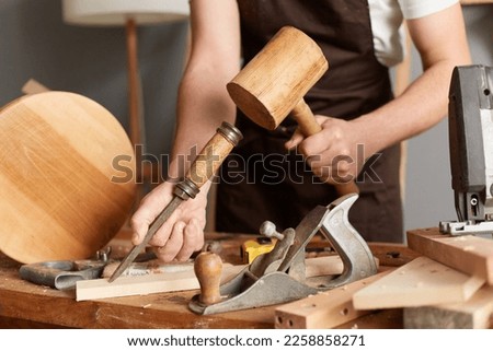 Closeup portrait of unknown faceless man master wearing brown apron using mallet and chisel for making wooden furniture, craftsman working in his joinery workshop.